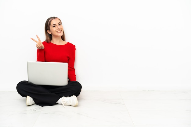 Junge kaukasische Frau mit einem Laptop, der lächelnd auf dem Boden sitzt und Siegeszeichen zeigt