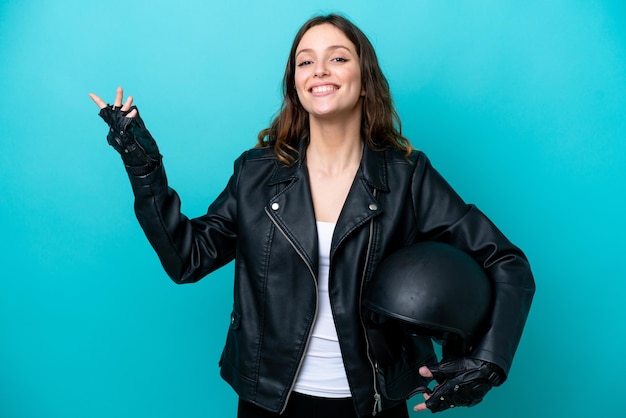 Junge kaukasische Frau mit einem Motorradhelm isoliert auf blauem Hintergrund, der die Hände zur Seite streckt, um zum Kommen einzuladen