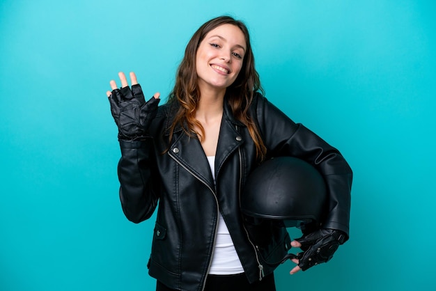 Junge kaukasische Frau mit einem Motorradhelm lokalisiert auf blauem Hintergrund, der mit der Hand mit glücklichem Ausdruck salutiert