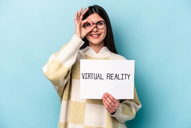 Junge kaukasische Frau mit einem Virtual-Reality-Plakat isoliert auf blauem Hintergrund aufgeregt, die Geste „OK“ im Auge zu behalten