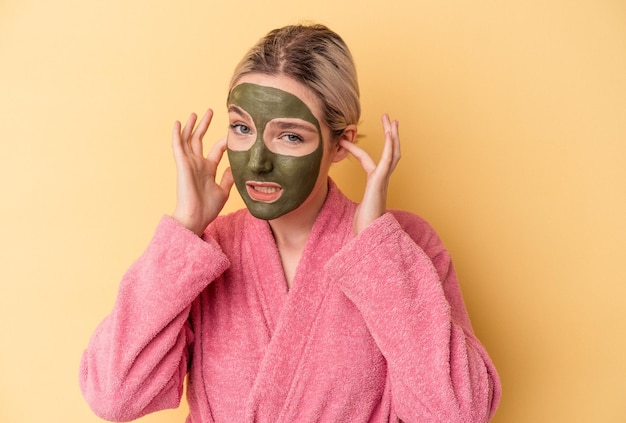 Junge kaukasische Frau mit Gesichtsmaske isoliert auf gelbem Hintergrund, die Ohren mit den Händen bedeckt.