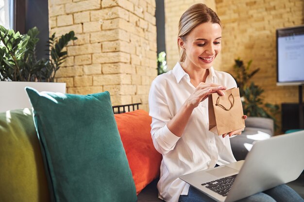 Junge kaukasische Frau mit grauem Laptop auf den Beinen, die Geschenk in den Händen hält