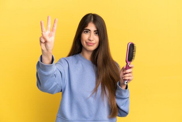 Junge kaukasische Frau mit Haarbürste isoliert auf blauem Hintergrund glücklich und zählt drei mit den Fingern