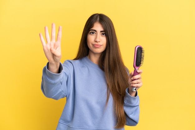 Junge kaukasische Frau mit Haarbürste isoliert auf blauem Hintergrund glücklich und zählt vier mit den Fingern