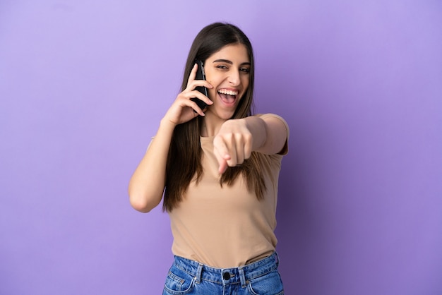 Junge kaukasische Frau mit Handy isoliert auf violettem Hintergrund und zeigt nach vorne mit glücklichem Ausdruck