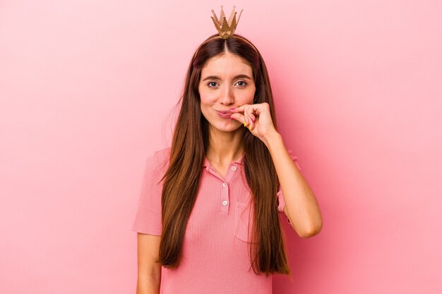 Junge kaukasische Frau mit Krone isoliert auf rosa Hintergrund mit Fingern auf den Lippen, die ein Geheimnis bewahren.