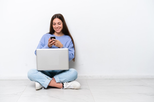 Junge kaukasische Frau mit Laptop sitzt auf dem Boden isoliert auf weißem Hintergrund und sendet eine Nachricht mit dem Handy