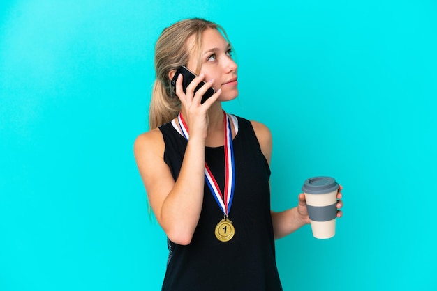 Foto junge kaukasische frau mit medaillen auf blauem hintergrund isoliert, die kaffee zum mitnehmen und ein handy hält