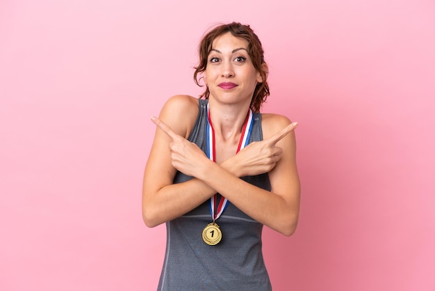 Junge kaukasische Frau mit Medaillen isoliert auf rosafarbenem Hintergrund, die auf die Seiten zeigen, die Zweifel haben