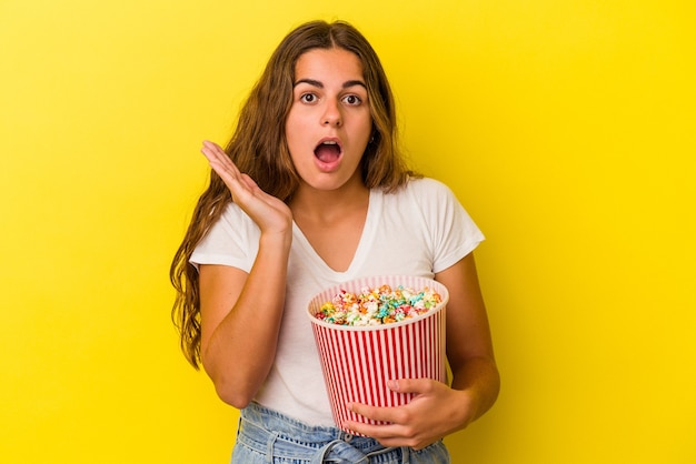 Junge kaukasische Frau mit Popcorn isoliert auf gelbem Hintergrund überrascht und schockiert.