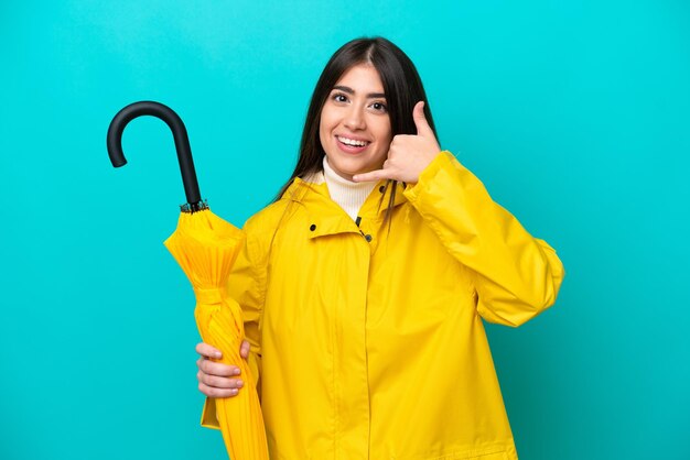 Junge kaukasische Frau mit regenfestem Mantel und Regenschirm isoliert auf blauem Hintergrund, die eine Telefongeste macht Ruf mich zurück Zeichen