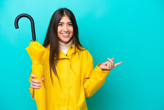 Junge kaukasische Frau mit regenfestem Mantel und Regenschirm isoliert auf blauem Hintergrund, die mit dem Finger zur Seite zeigt