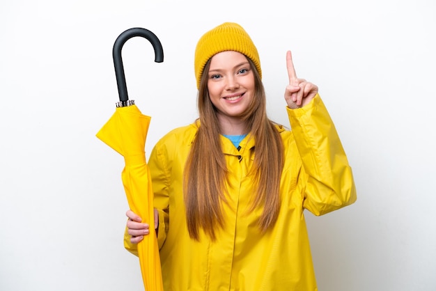 Junge kaukasische Frau mit regenfestem Mantel und Regenschirm isoliert auf weißem Hintergrund, die auf eine großartige Idee zeigt