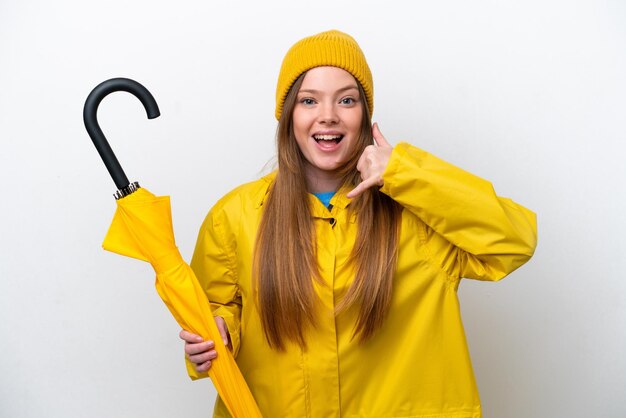 Junge kaukasische Frau mit regenfestem Mantel und Regenschirm isoliert auf weißem Hintergrund, die eine Telefongeste macht Ruf mich zurück Zeichen