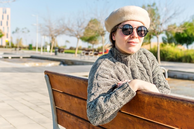 Junge kaukasische Frau mit runder Sonnenbrille und Baskenmütze, die an einem sonnigen Tag in einem Park in die Kamera blickt