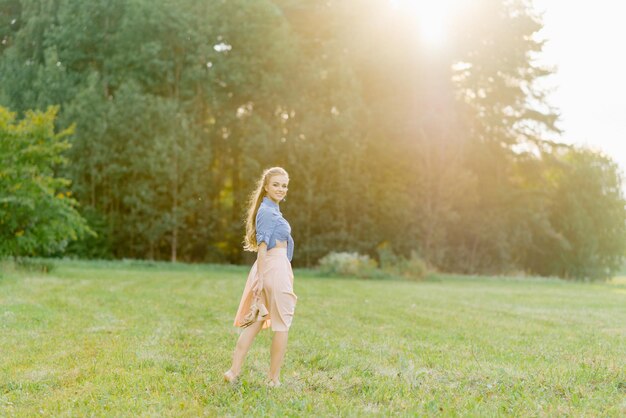 Junge kaukasische Frau oder Mädchen in einem Sommerkleid und einem Jeanshemd geht auf dem Gras und hält Schuhe in ihren Händen, die sich an einem Wochenende im Sommer außerhalb der Stadt entspannen