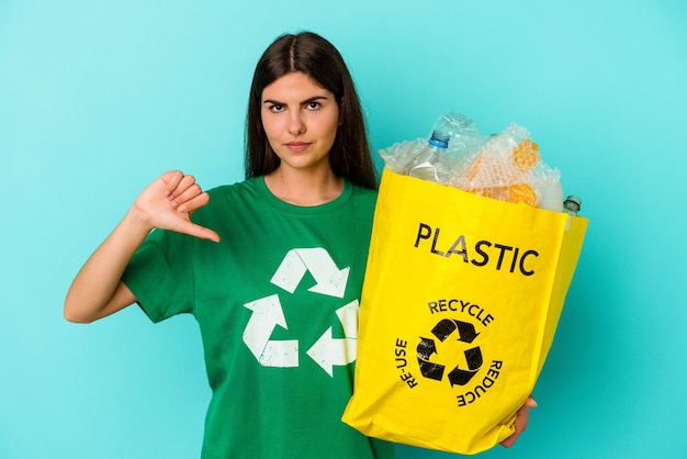 Junge kaukasische Frau recycelte Plastik lokalisiert auf blauem Hintergrund, der eine Abneigungsgeste zeigt, Daumen nach unten. Uneinigkeit Konzept.