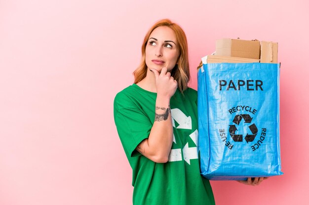 Junge kaukasische Frau recyceltes Papier einzeln auf rosafarbenem Hintergrund, das seitlich mit zweifelhaftem und skeptischem Ausdruck schaut.