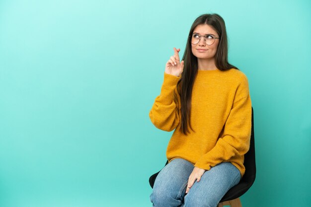 Junge kaukasische Frau sitzt auf einem Stuhl isoliert auf blauem Hintergrund mit gekreuzten Fingern und wünscht das Beste