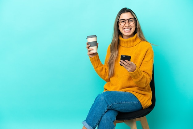 Junge kaukasische Frau sitzt auf einem Stuhl isoliert auf blauem Hintergrund und hält Kaffee zum Mitnehmen und ein Handy a
