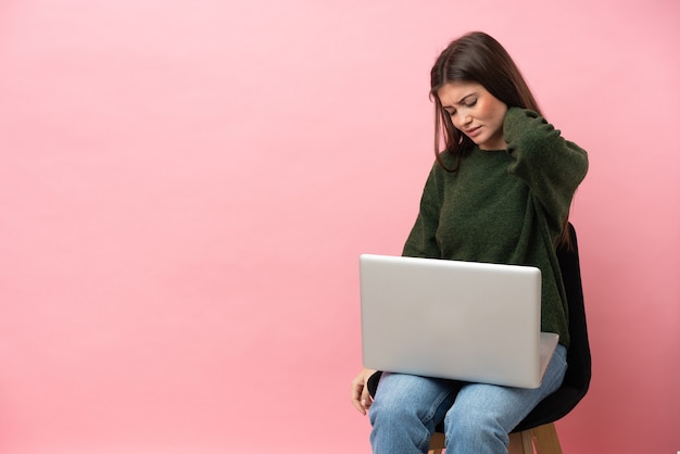 Junge kaukasische Frau sitzt auf einem Stuhl mit ihrem Laptop auf rosafarbenem Hintergrund mit Nackenschmerzen