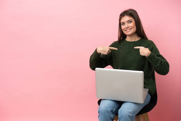 Junge kaukasische Frau sitzt auf einem Stuhl mit ihrem Laptop isoliert auf rosa Hintergrund stolz und selbstzufrieden