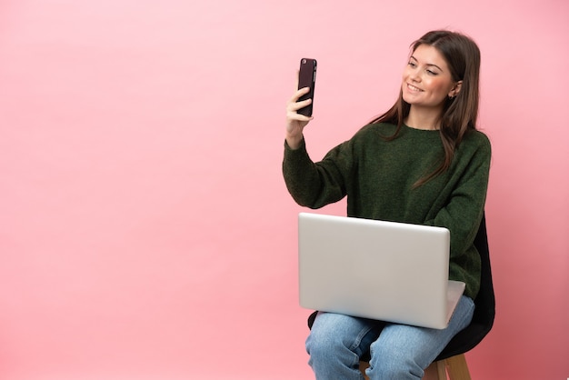 Junge kaukasische Frau sitzt auf einem Stuhl mit ihrem Laptop isoliert auf rosa Hintergrund und macht ein Selfie