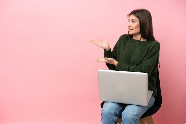 Junge kaukasische Frau sitzt auf einem Stuhl mit ihrem Laptop isoliert auf rosafarbenem Hintergrund mit überraschtem Gesichtsausdruck