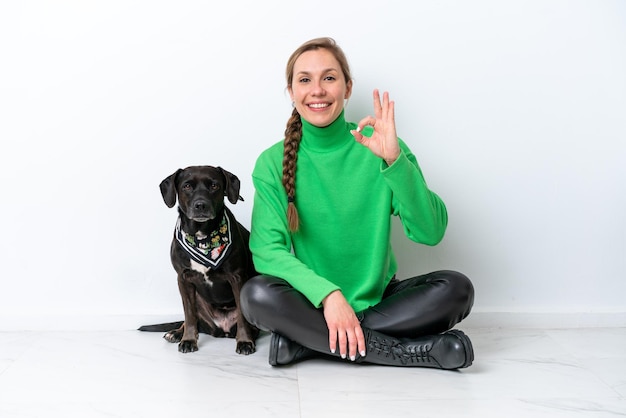 Foto junge kaukasische frau sitzt mit seinem welpen isoliert auf weißem hintergrund auf dem boden und zeigt mit den fingern ein ok-zeichen