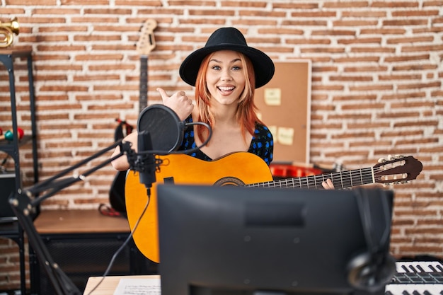 Junge kaukasische Frau spielt klassische Gitarre im Musikstudio und zeigt mit dem Daumen nach oben zur Seite und lächelt glücklich mit offenem Mund