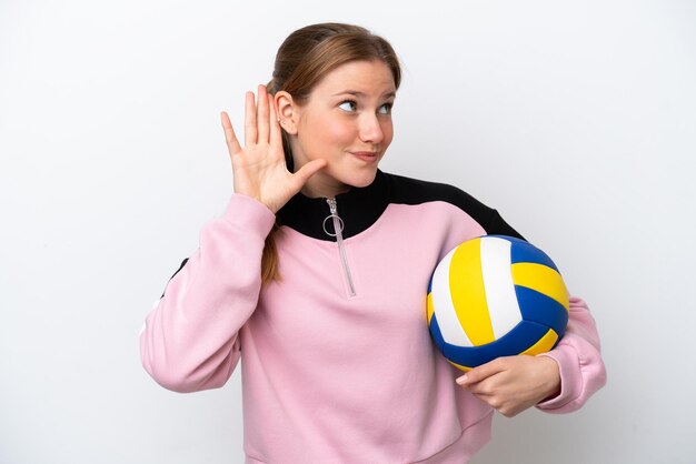 Junge kaukasische Frau spielt Volleyball isoliert auf weißem Hintergrund und hört etwas, indem sie die Hand auf das Ohr legt