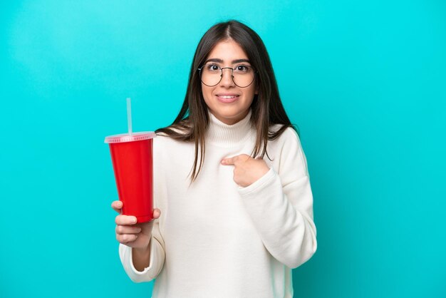 Junge kaukasische Frau trinkt Soda isoliert auf blauem Hintergrund mit überraschendem Gesichtsausdruck