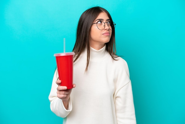 Junge kaukasische Frau trinkt Soda isoliert auf blauem Hintergrund und blickt nach oben