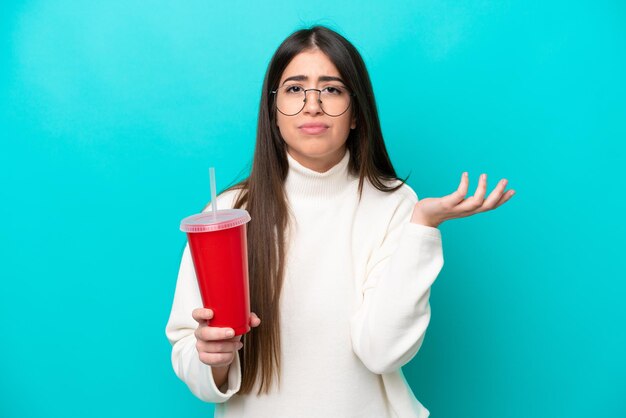 Junge kaukasische Frau trinkt Soda isoliert auf blauem Hintergrund und hat Zweifel, während sie die Hände hebt