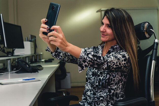 Junge kaukasische Geschäftsfrau, die lachend sitzt und ein Selfie im Büro macht