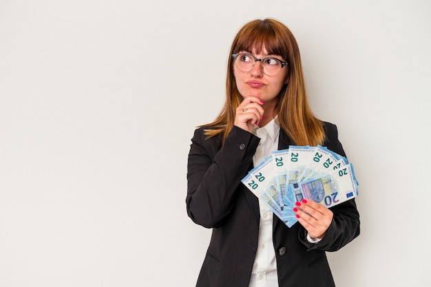 Junge kaukasische Geschäftsfrau, die Rechnungen lokalisiert auf weißem Hintergrund hält, der seitlich mit zweifelhaftem und skeptischem Ausdruck schaut.