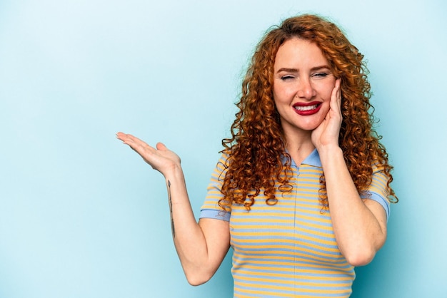 Junge kaukasische Ingwerfrau, die auf blauem Hintergrund isoliert ist, hält Kopierbereich auf einer Handfläche, hält die Hand über die Wange. Erstaunt und erfreut