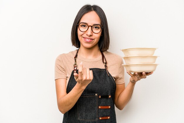 Junge kaukasische Kellnerin, die Tupperware isoliert auf weißem Hintergrund hält und mit dem Finger auf Sie zeigt, als würde sie einladen, näher zu kommen