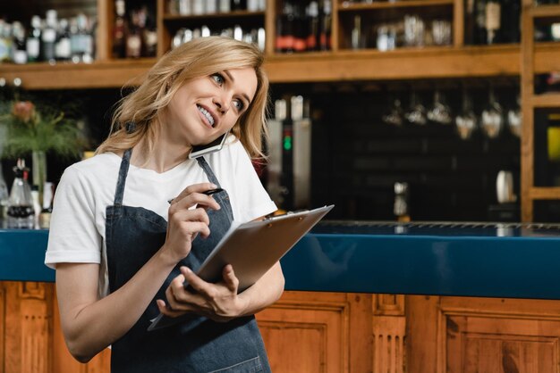 Foto junge kaukasische kellnerin, kleinunternehmerin, barkeeper, barista, schreibt bestellungen in eine schreibtafel, trägt eine blaue schürze, spricht am telefon für take-out-take-away-lieferung, schaut in die kamera an der bar.