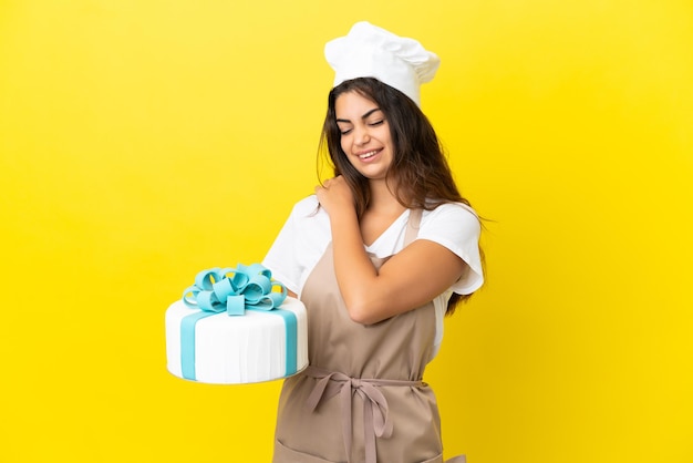 Junge kaukasische Konditorfrau mit einem großen Kuchen einzeln auf gelbem Hintergrund, der unter Schmerzen in der Schulter leidet, weil sie sich angestrengt hat