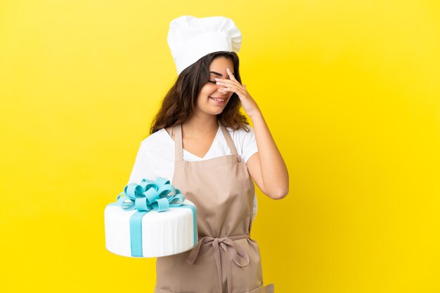 Junge kaukasische Konditorin mit einem großen Kuchen isoliert auf gelbem Hintergrund lachend