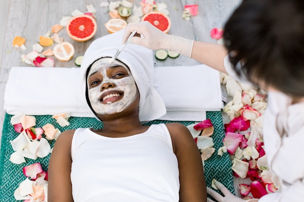 Junge kaukasische Kosmetikerin, die kosmetische Maske auf dem Gesicht der afrikanischen Frau in einem Schönheitssalon anwendet. Frau mit Maske auf dem Gesicht entspannend in einem Schönheitssalon