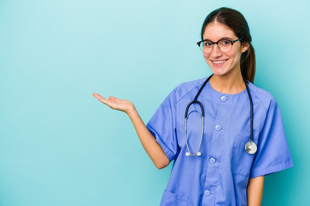 Junge kaukasische Krankenschwester auf blauem Hintergrund isoliert, die einen Kopienraum auf einer Handfläche zeigt und eine andere Hand an der Taille hält.