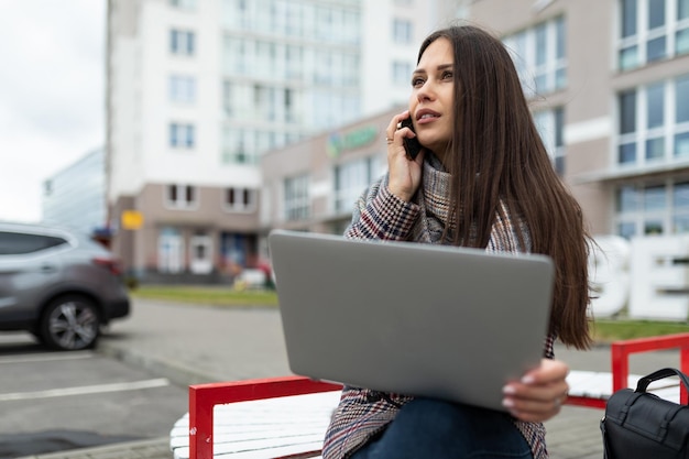 Junge kaukasische Managerin telefoniert mit einem Laptop auf den Knien und sitzt auf einer Bank