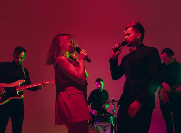 Junge kaukasische Musikerband, die im Neonlicht auf rotem Studiohintergrund aufführt
