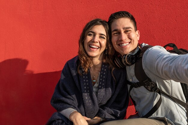 Junge kaukasische Paare machen ein Selfie-Foto lachen. Mann und Frau mit Freizeitkleidung, Reisenden oder Studenten. Rote Wand im Hintergrund.