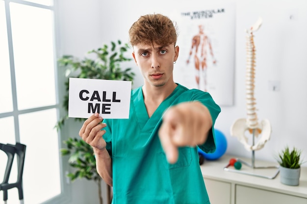Junge kaukasische Physio Mann hält rufen Sie mich Banner in der Klinik zeigt mit dem Finger auf die Kamera und zu Ihnen zuversichtliche Geste sieht ernst