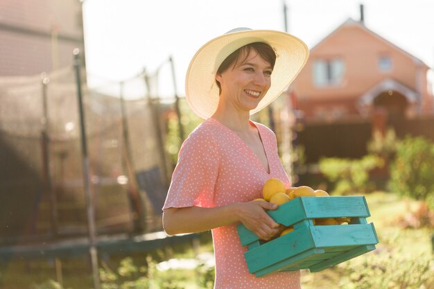 Junge kaukasische positive Gärtnerin in Hut und rosa Kleid hält an einem sonnigen Sommertag eine Kiste mit Zitronen in den Händen. Konzept der Landwirtschaft und des biologischen Gartens