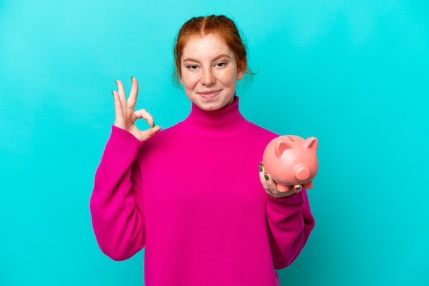 Foto junge kaukasische rötliche frau, die ein sparschwein isoliert auf blauem hintergrund hält und mit den fingern ein ok-zeichen zeigt