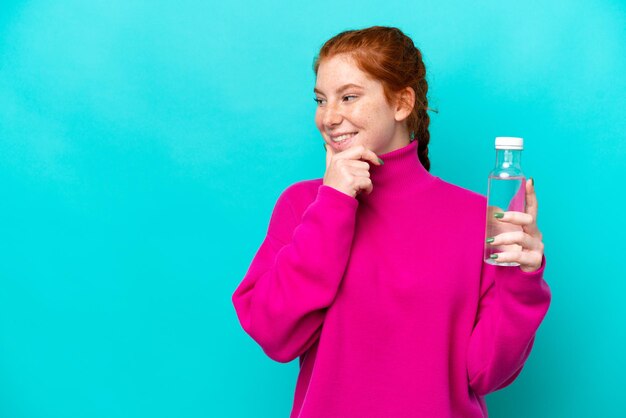 Foto junge kaukasische rötliche frau mit einer flasche wasser isoliert auf blauem hintergrund, die eine idee denkt und zur seite schaut